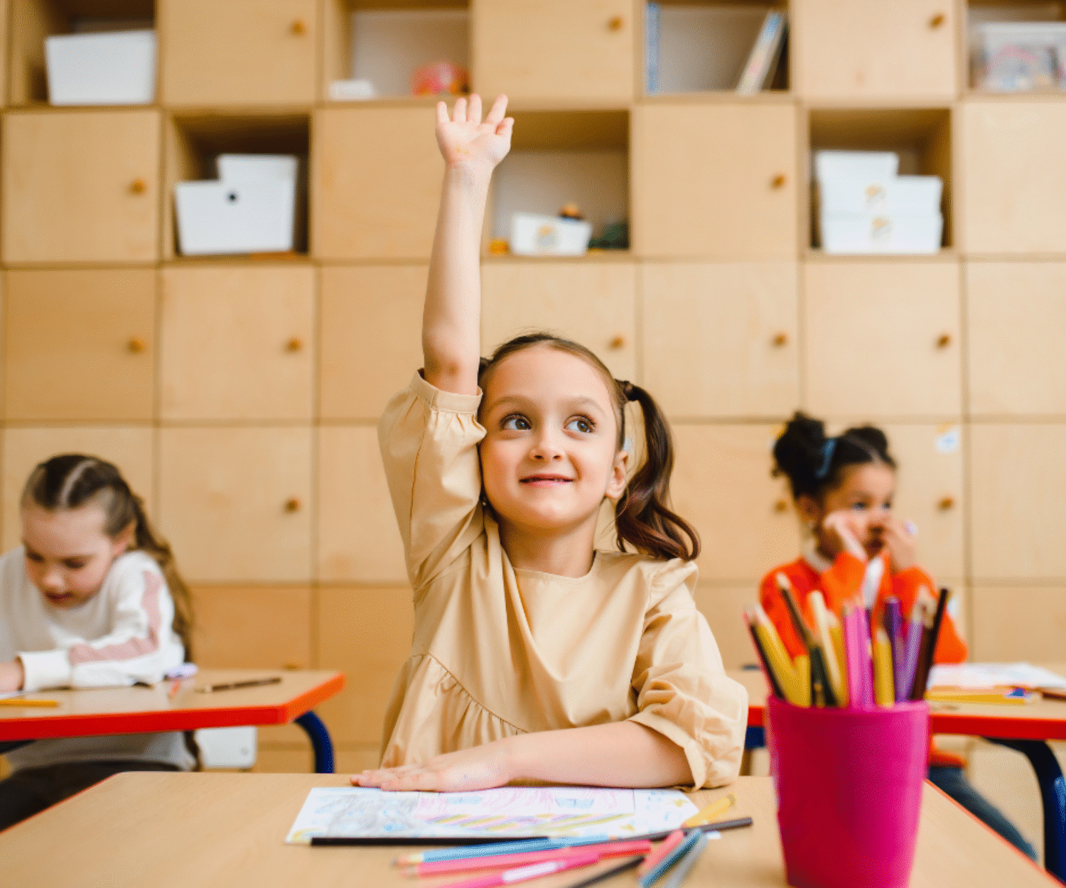 Photo d'une petite filles souriante avec des couettes qui lève la main en classe. Elle est dans une école avec d'autres enfants et elle fait un coloriages avec des crayons de couleur dans un pot rose. Il y a des étagères derrière.