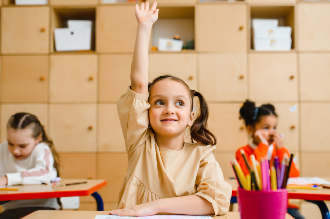 Photo d'une petite filles souriante avec des couettes qui lève la main en classe. Elle est dans une école avec d'autres enfants et elle fait un coloriages avec des crayons de couleur dans un pot rose. Il y a des étagères derrière.