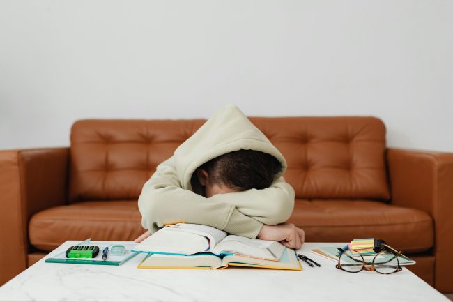 Enfant qui boude devant ses devoirs. Il a une capuche sur la tête.
